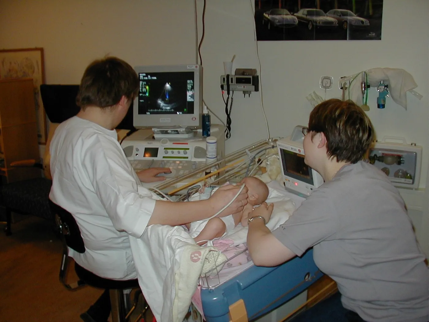 Een echografist voert een pediatrische echocardiografie (hartecho) uit bij een baby met een medisch echografieapparaat met digitaal beeldscherm aan het bed, terwijl de moeder van de baby aanwezig is.