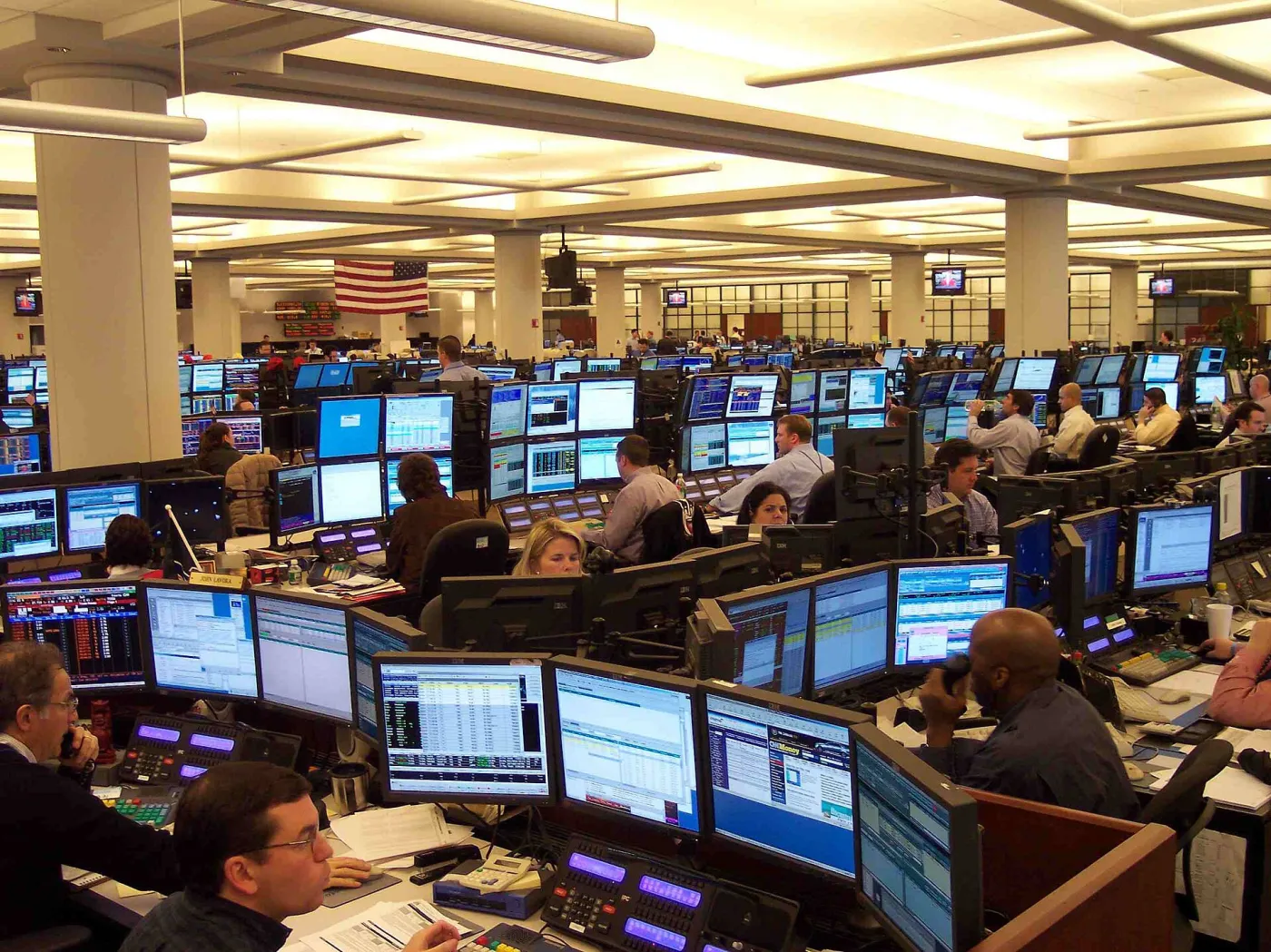 Een drukke handelsvloer in een oliehandelskantoor in Houston, met rijen handelaren aan bureaus vol computerschermen die actuele marktgegevens en grafieken tonen, met een Amerikaanse vlag op de achtergrond.
